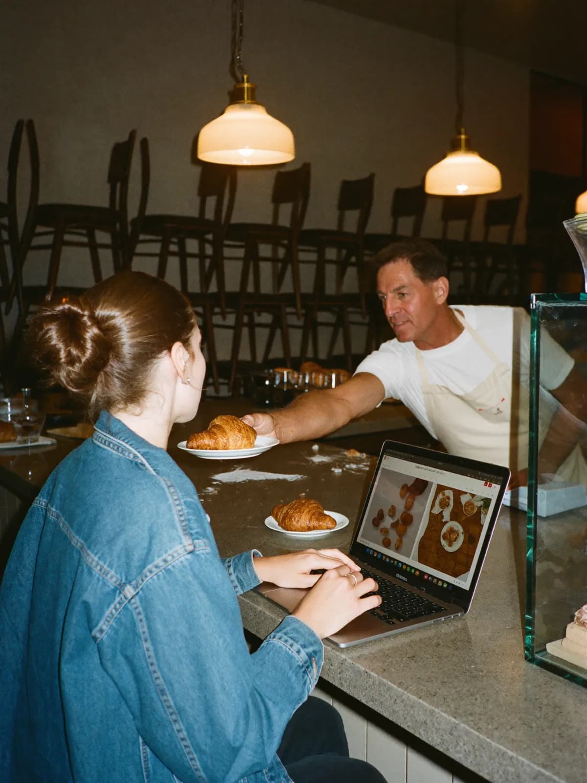 After closing, a freelancer builds the bakery’s website at the counter while the baker passes her a croissant