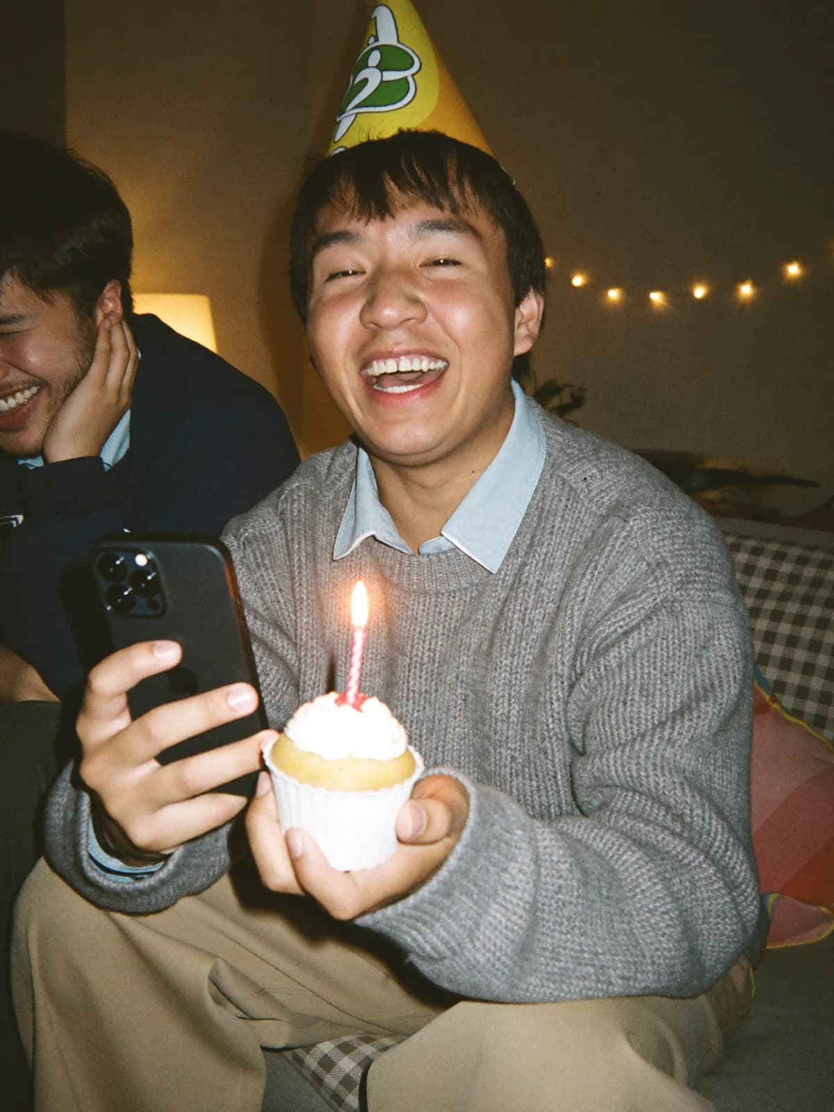 A young man in a party hat belts happy birthday into a video call, holding up a cupcake with one candle, while his roommate laughs into his hand