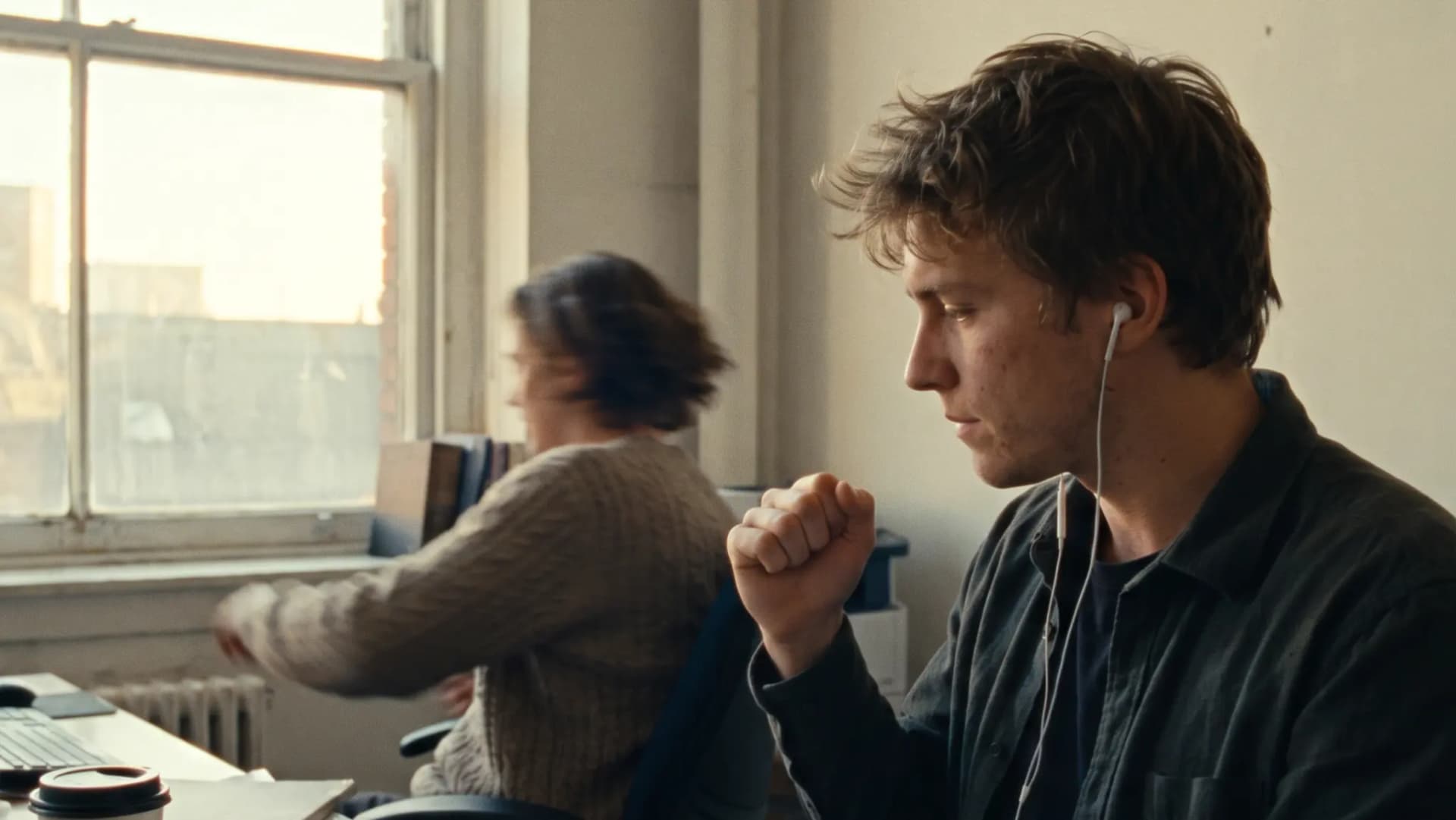 A young account executive in wired earphones does a tiny, restrained victory fist-pump at a window desk as a call ends, a colleague working behind him