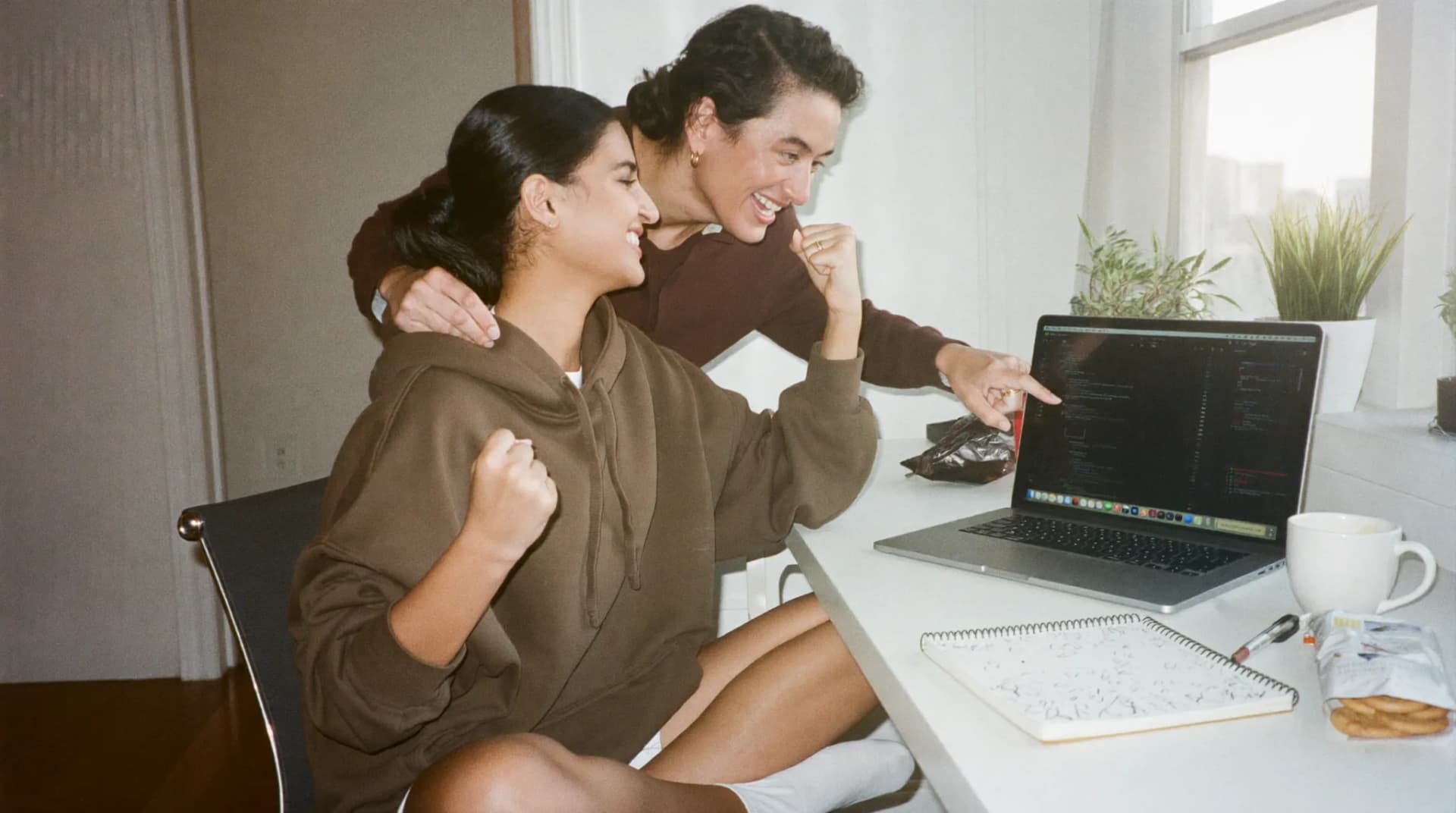 An intern in an oversized hoodie pumps both fists at her laptop as her first code is approved, while a teammate leaning over her shoulder laughs and points at the screen, chai and biscuits on the desk