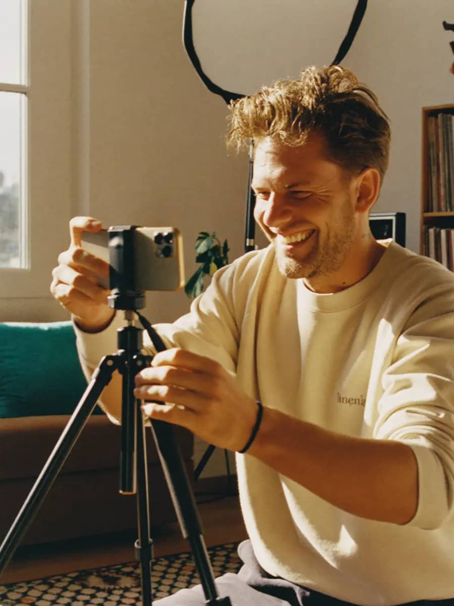 A content creator grinning at the framing on a phone clamped to a tripod.