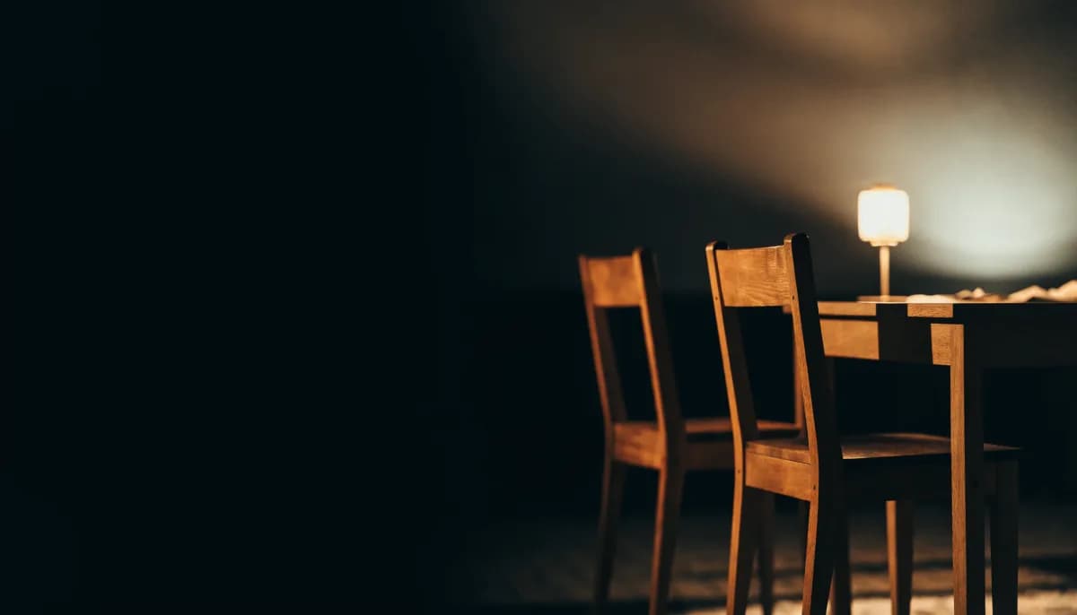 A brass desk lamp lit over a bare dark wooden desk late at night, the room falling away into shadow.